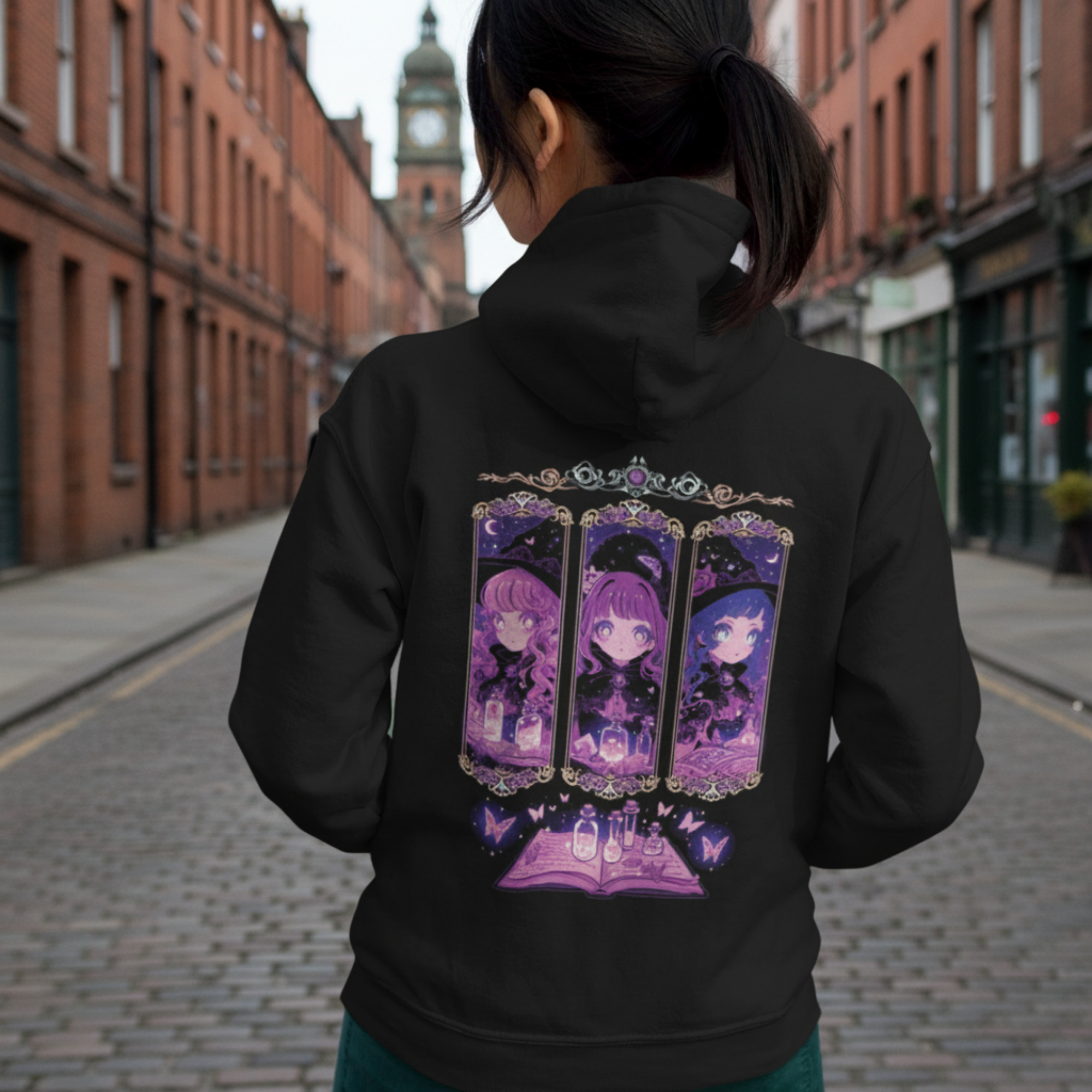 Woman in a black Coven Sisters Kawaii Witch Hoodie standing on a cobbled street, back turned to show the kawaii witch triptych and glowing grimoire design.