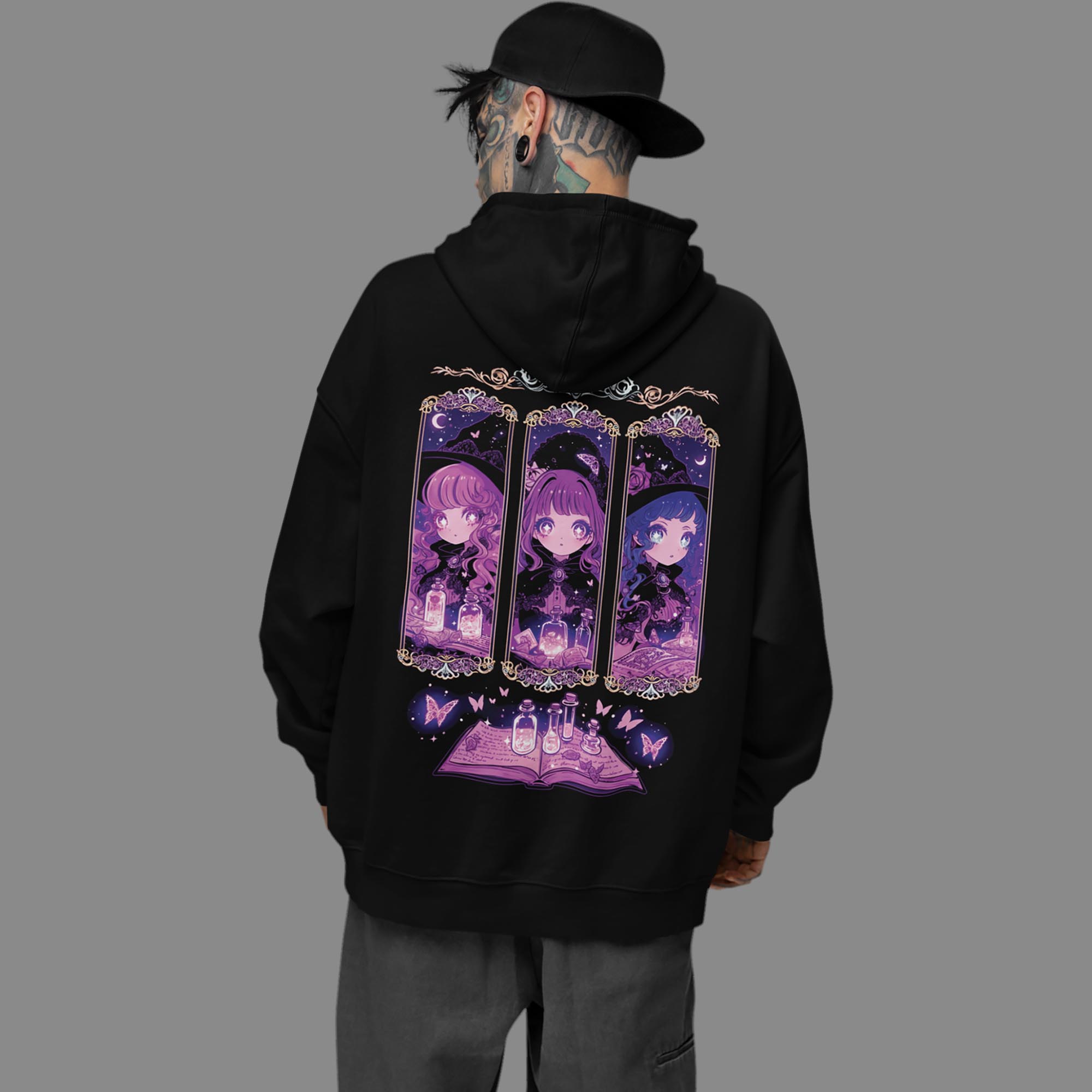 Tattooed man wearing an oversized black Coven Sisters Kawaii Witch Hoodie, turned to show the large kawaii witch coven and grimoire back print.